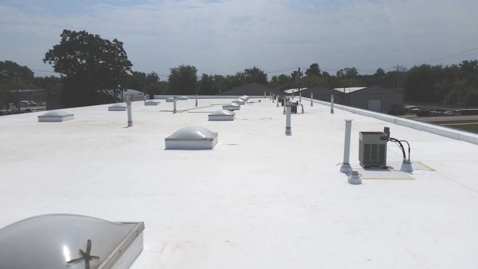 White commercial roof with skylights, vents, and HVAC units, set against a cloudy sky and trees.