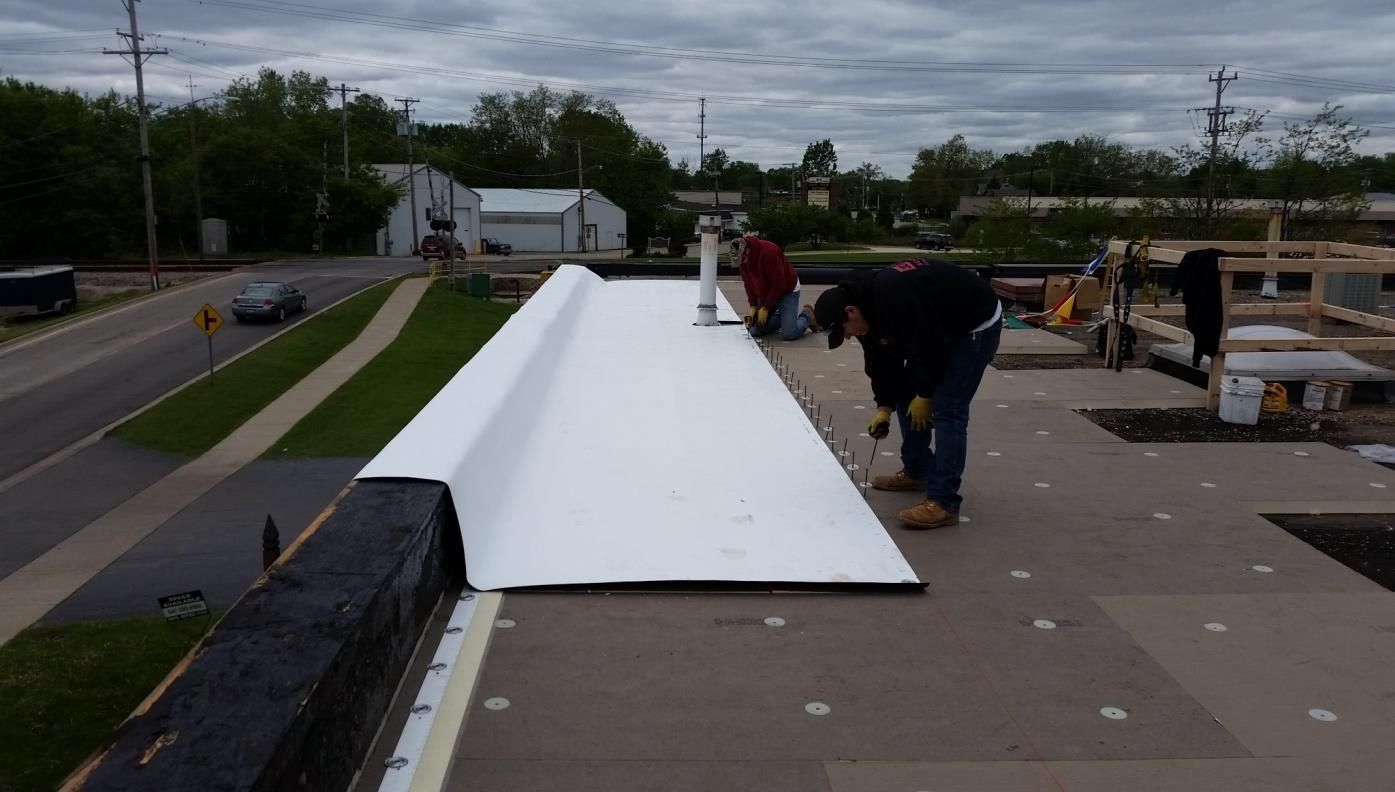 Roofers installing white roofing material on a flat commercial roof; one kneeling, two standing, outdoor setting.