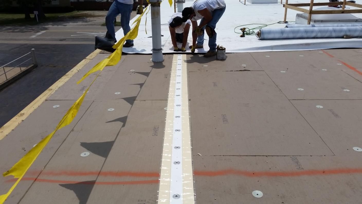 Workers installing roofing material on a building's flat roof, using tools and measuring equipment.