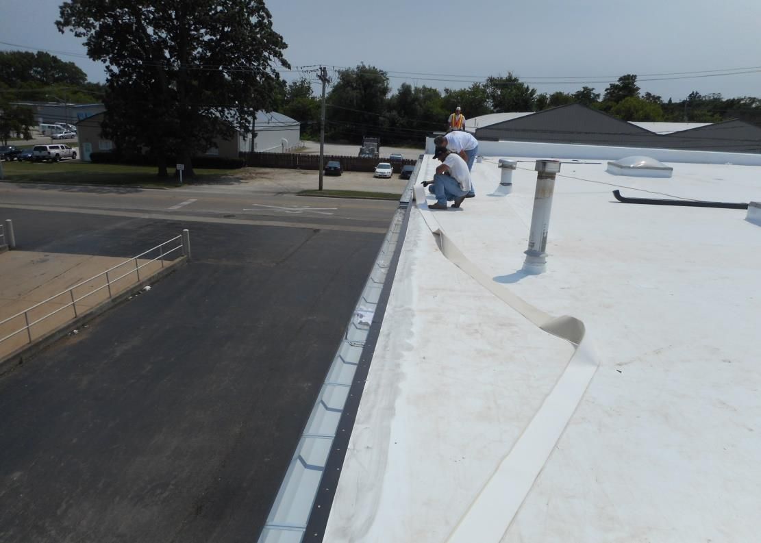 Two workers on a white commercial roof; one kneels, the other stands. A gutter and street are visible.