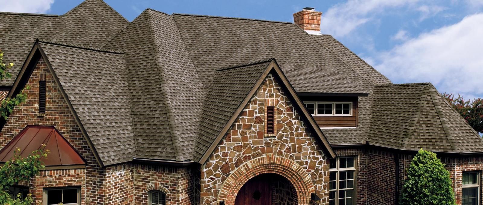 Stone house with a brown shingle roof, chimney, and arched entryway, against a blue sky.