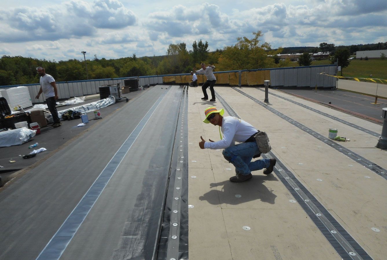 Roofing crew installing a flat roof. Man kneels, giving thumbs up. Others work nearby, sunny day.