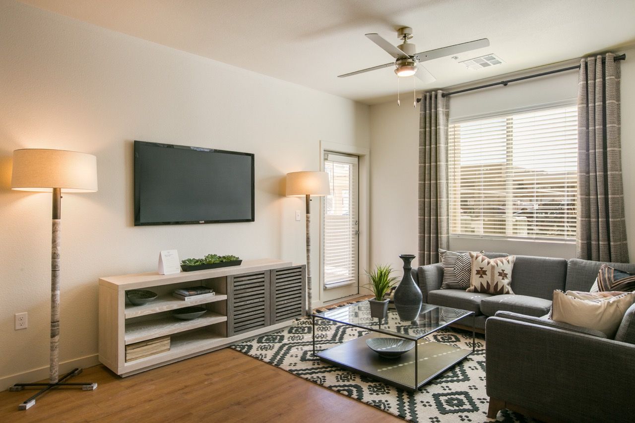 Living room in an apartment with a grey sectional sofa, wall-mounted TV, and a large window with blinds.