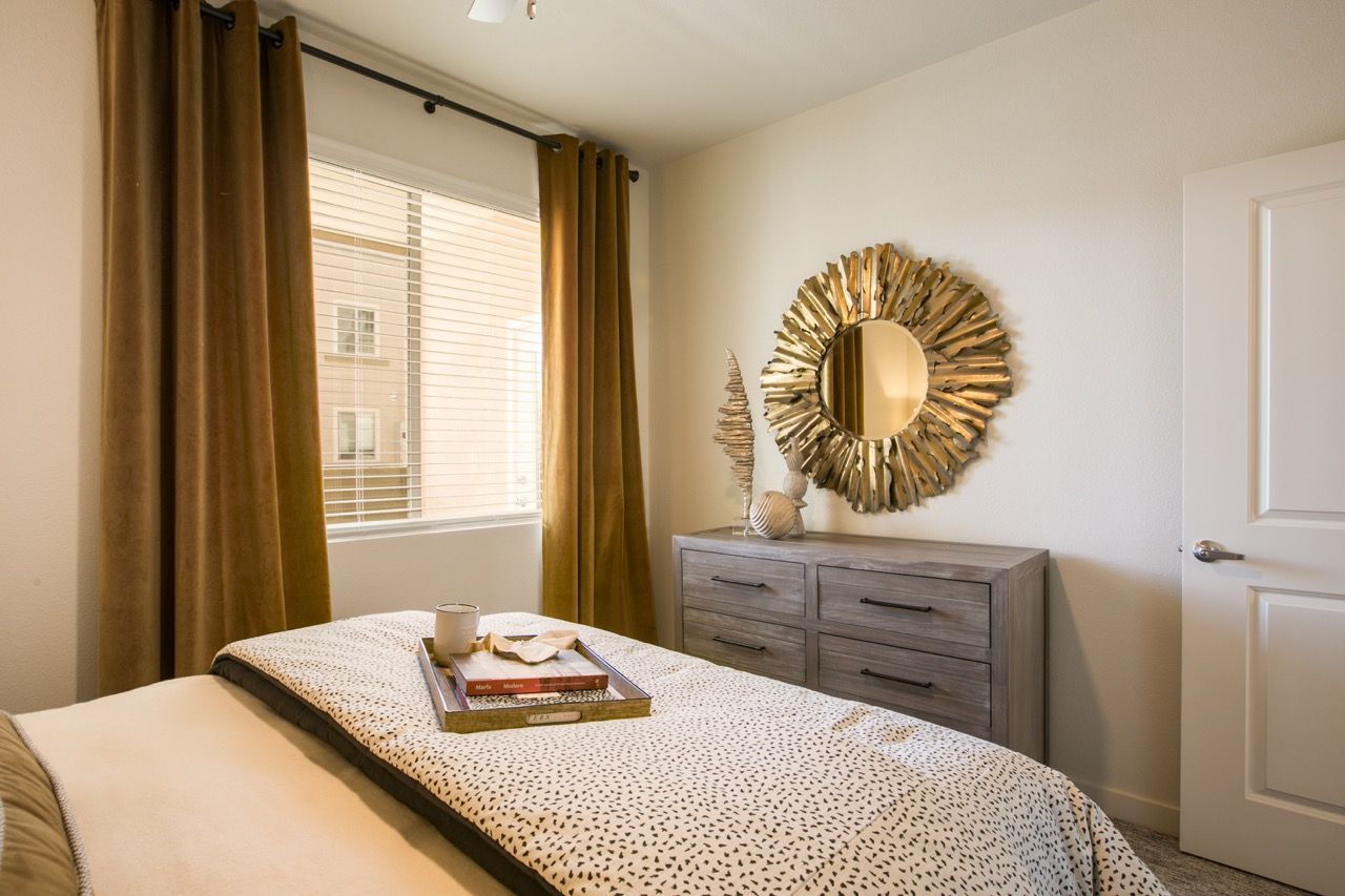 Bedroom with bed, gray dresser, sunburst mirror, and window with brown curtains.