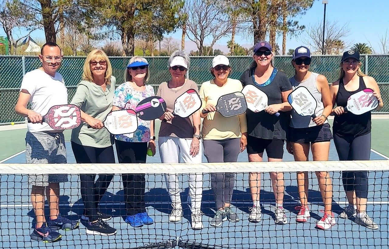A group poses behind a pickleball net with their paddles raised to chest height.