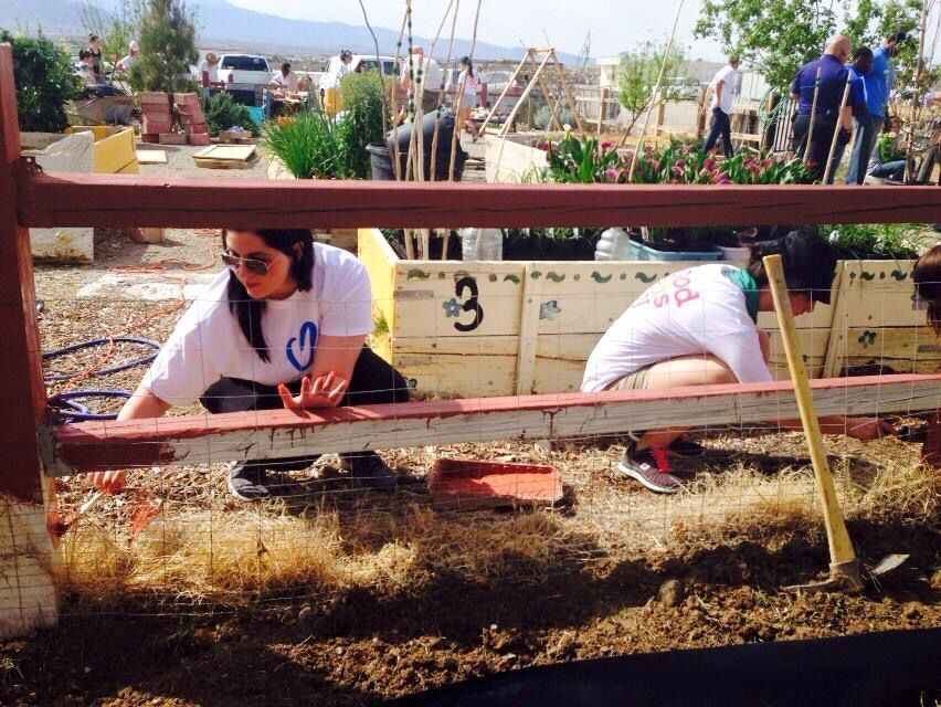 Two volunteers in white shirts are crouched down behind a half-painted wooden fence. 