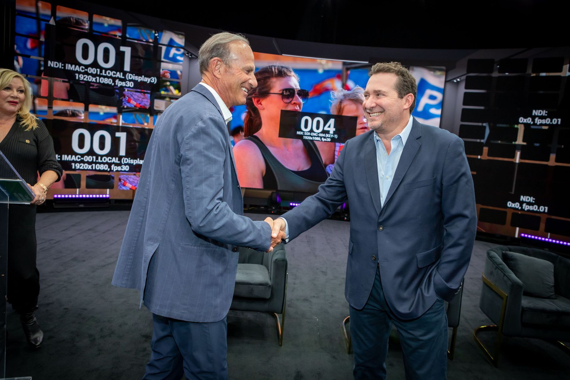Two smiling individuals in dark blue suits shake hands in front of an interview set stage with video monitors