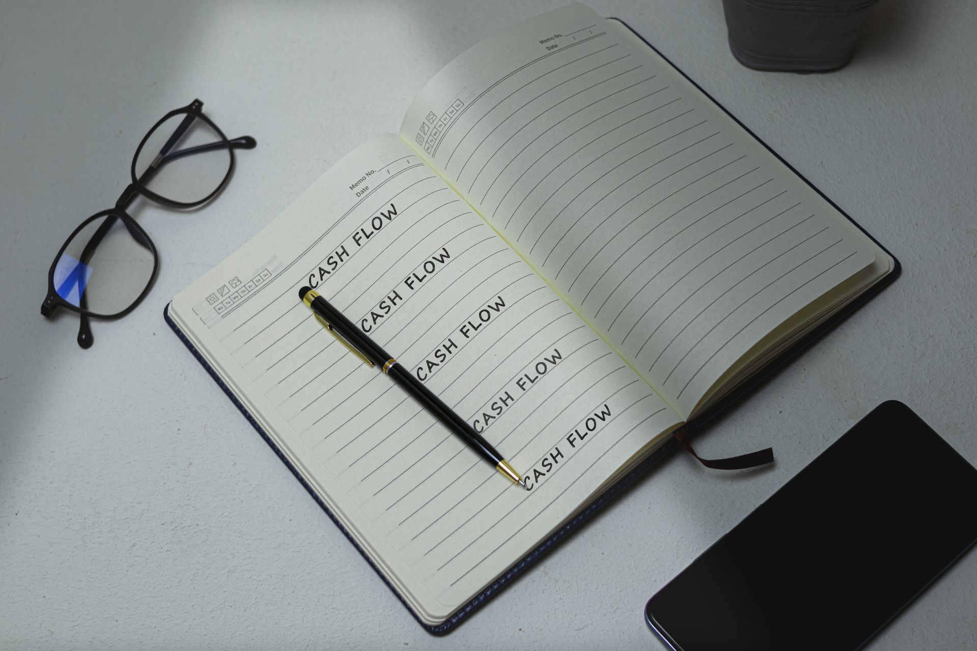 Open notebook with the words cash flow repeated on the page beside glasses and a pen.