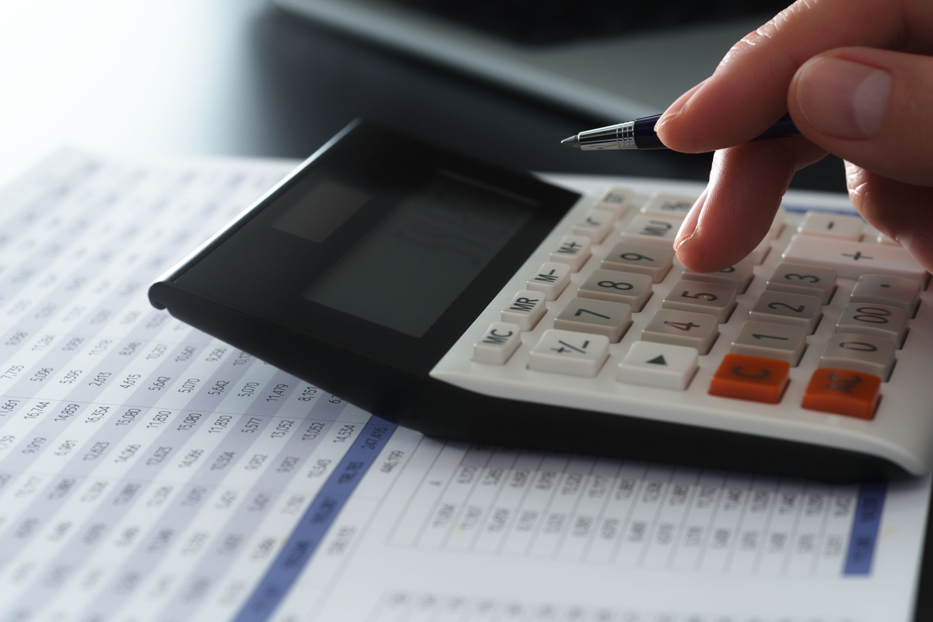 A hand using a white calculator with orange and gray buttons over a printed financial spreadsheet. A hand using a white calculator with orange and gray buttons over a printed financial spreadsheet.