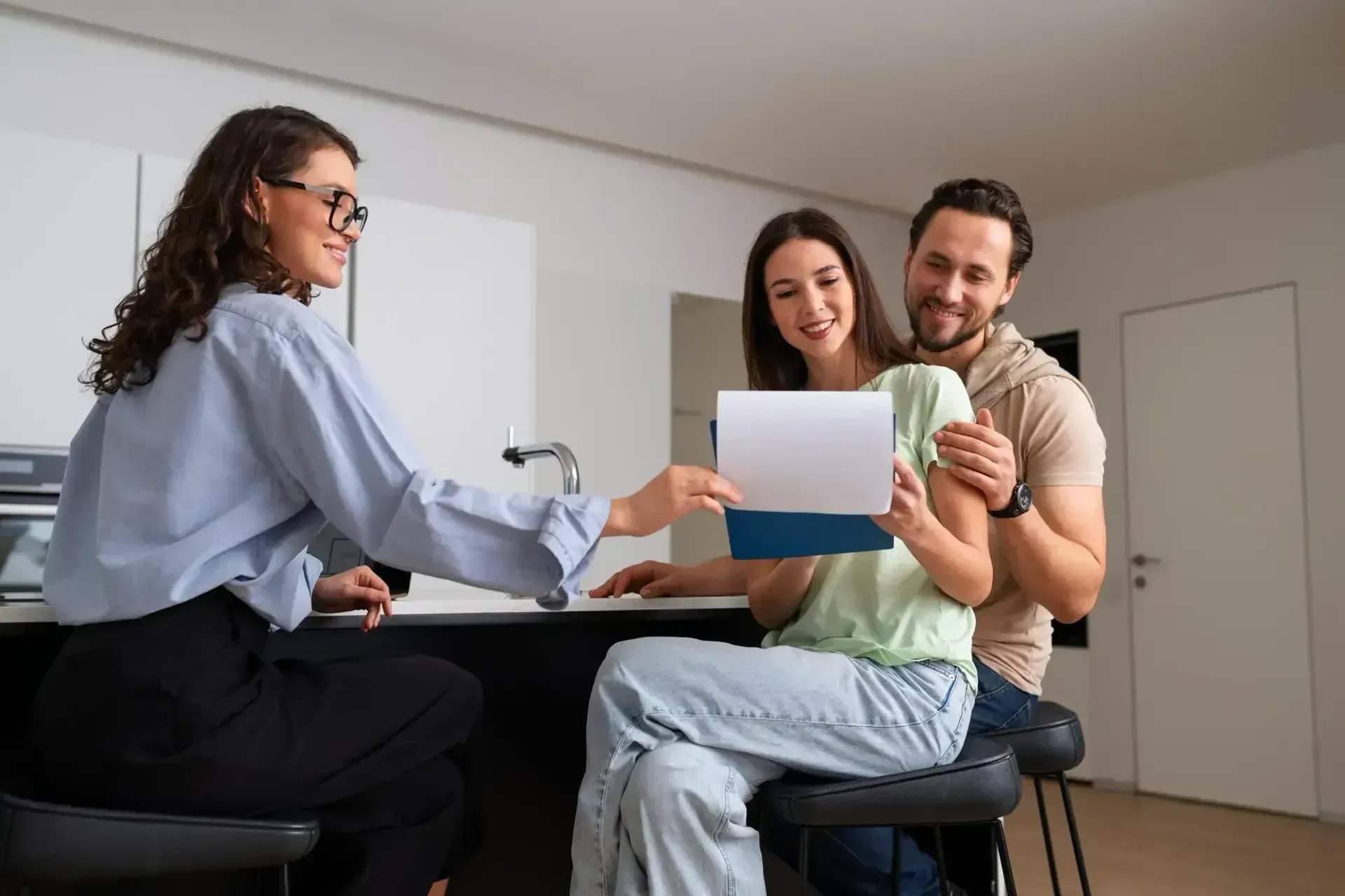 Mulher de óculos entrega papelada a um casal sorridente, possivelmente uma transação imobiliária.