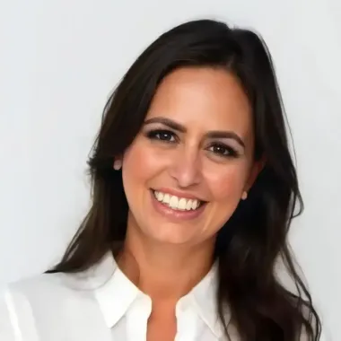 Mulher de cabelos escuros sorrindo, vestindo uma camisa branca, contra um fundo claro.