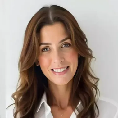 Mulher de cabelos castanhos sorri, vestindo uma camisa branca, sobre um fundo branco.