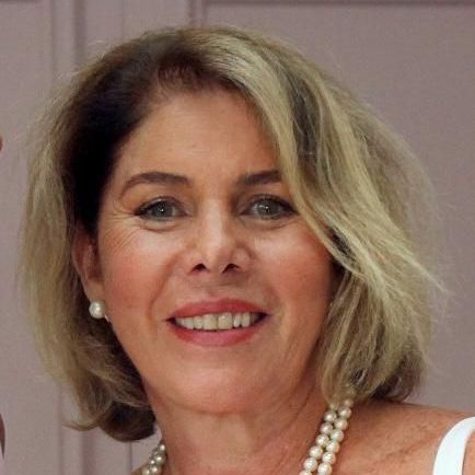 Mulher de pele clara e cabelo loiro curto, usando um colar de pérolas, sorri e está dentro de casa.