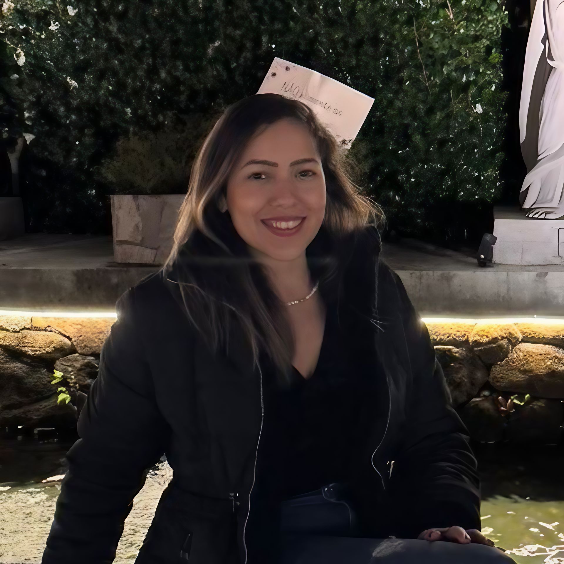 Mulher sorrindo com uma jaqueta preta ao ar livre à noite, com árvores e uma estátua ao fundo.