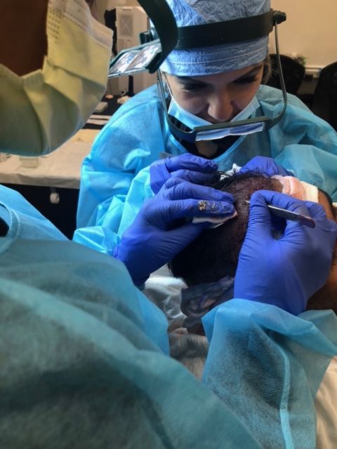 A medical professional wearing blue scrubs and a head-mounted light performs a procedure on a patient's scalp.