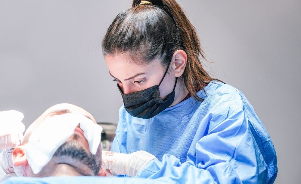 Surgeon wearing mask, blue scrubs, and gloves tending to a patient's head during a procedure.