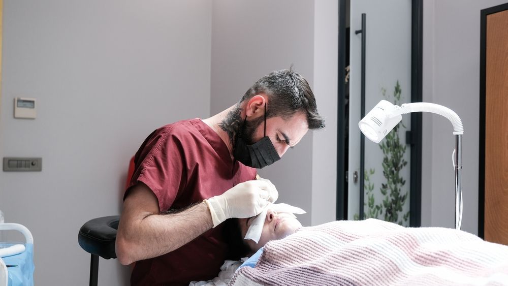 Person in maroon scrubs and mask performing a procedure on a person's face.  Medical setting, soft lighting.