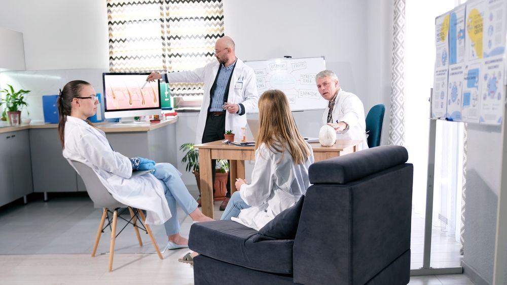 Doctors in lab coats, discussing a presentation displayed on a computer screen, in an office.
