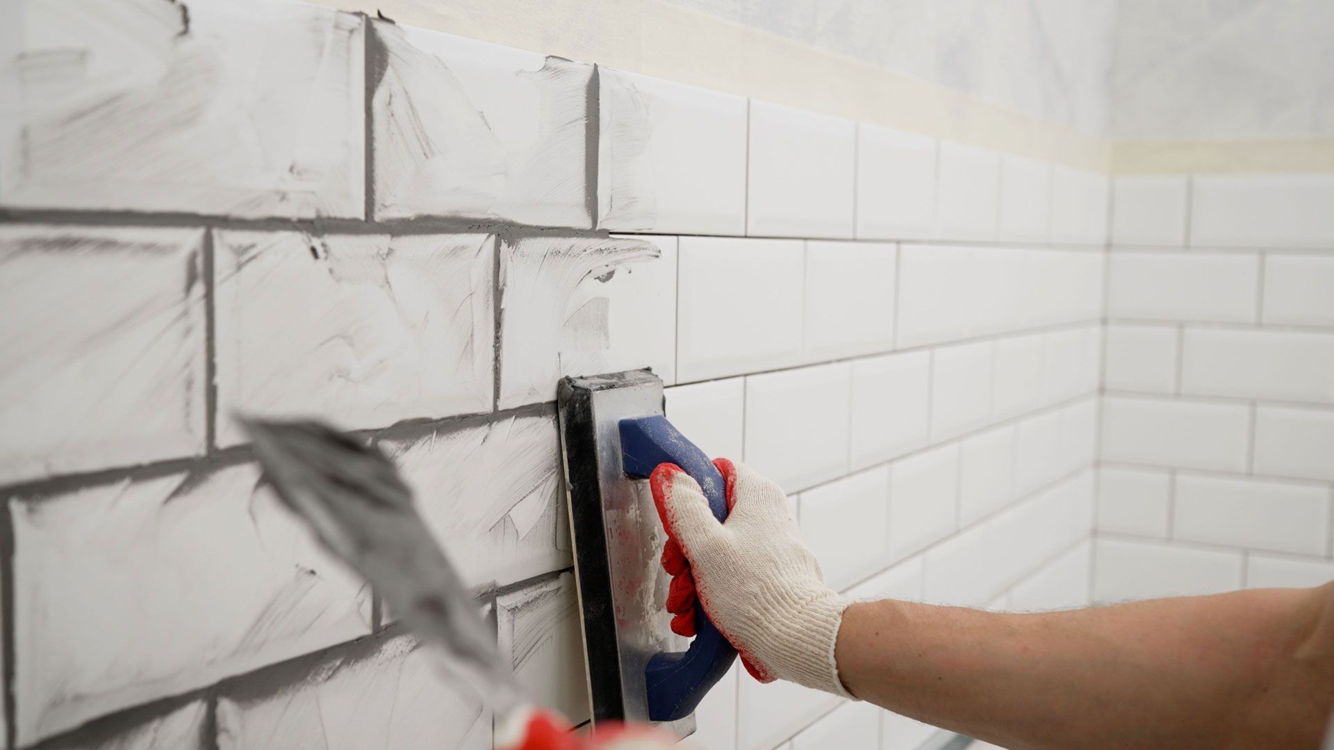 Person applying gray grout to white subway tiles on a wall with a trowel.