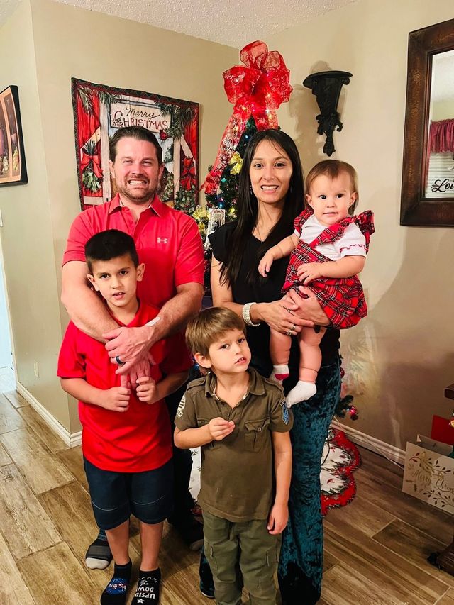 Family of five poses near a Christmas tree. Adults hold children; all smile.