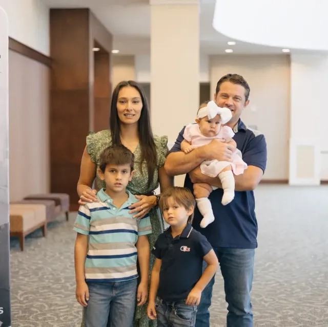 Family of five posing together; woman, man holding baby, and two boys. Indoor setting.