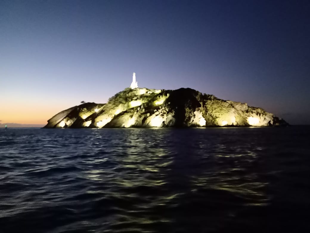 Una isla de noche, iluminada por luces. Una estructura, posiblemente un faro, se alza sobre la isla rocosa, reflejándose en las aguas oscuras.