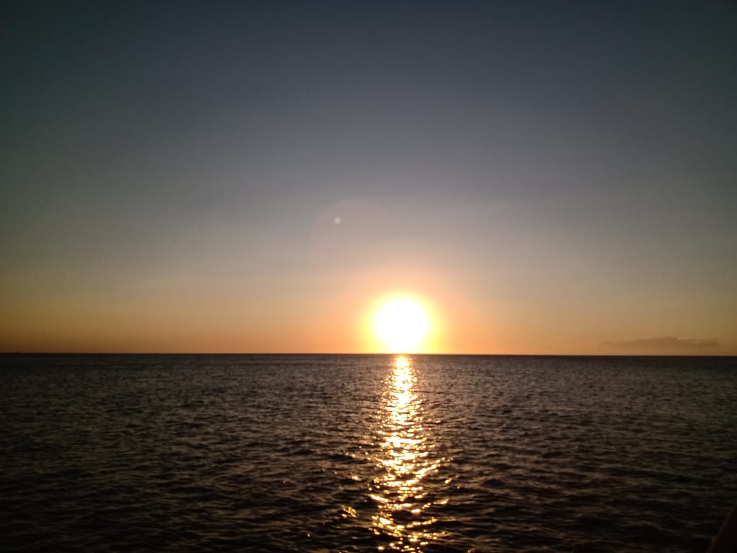Atardecer sobre el océano. El sol es un orbe naranja brillante que se refleja en el agua oscura, con un cielo degradado de naranja a azul.