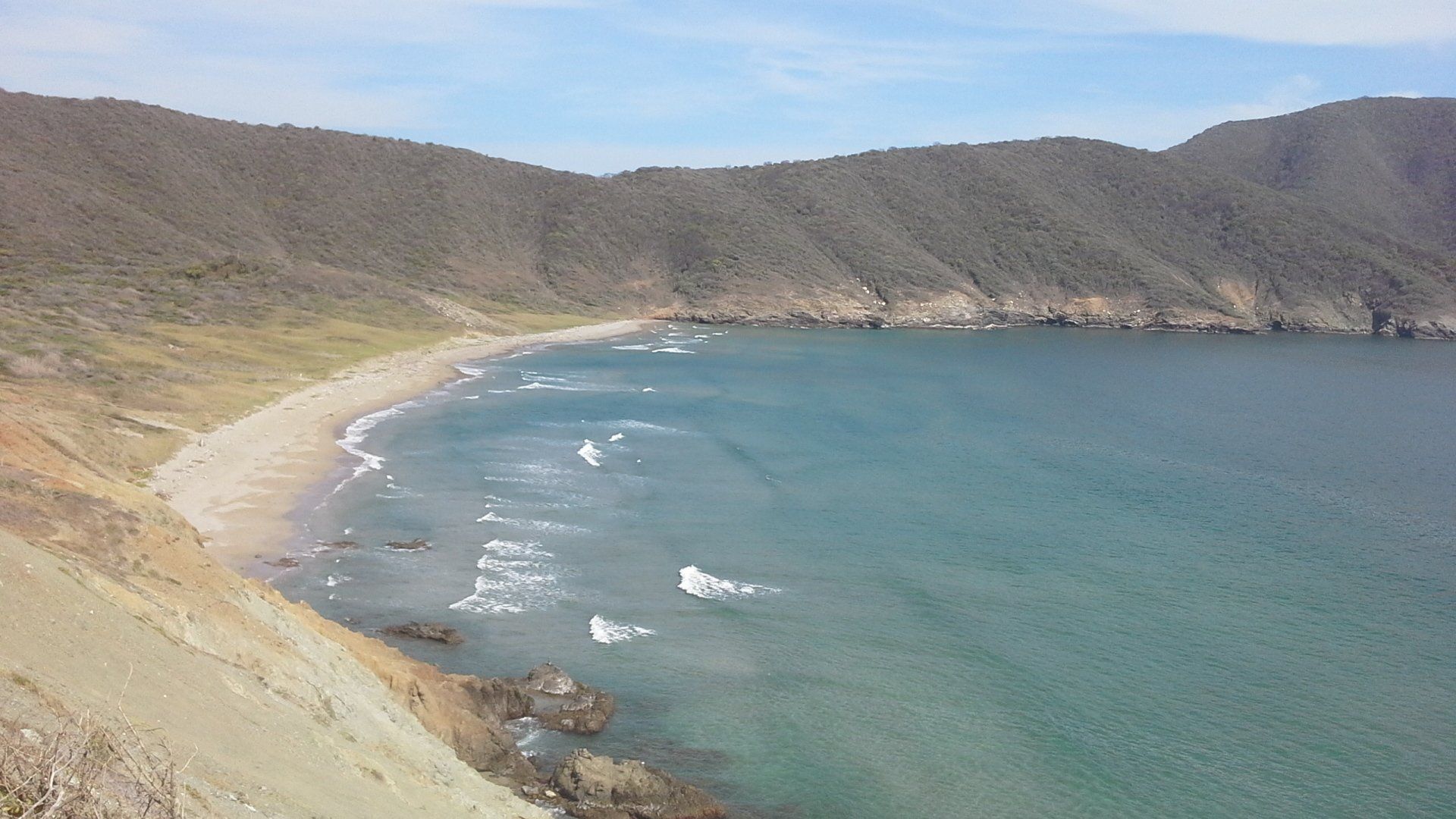 Vista costera de una playa de arena con olas, enclavada entre colinas bajo un cielo azul.