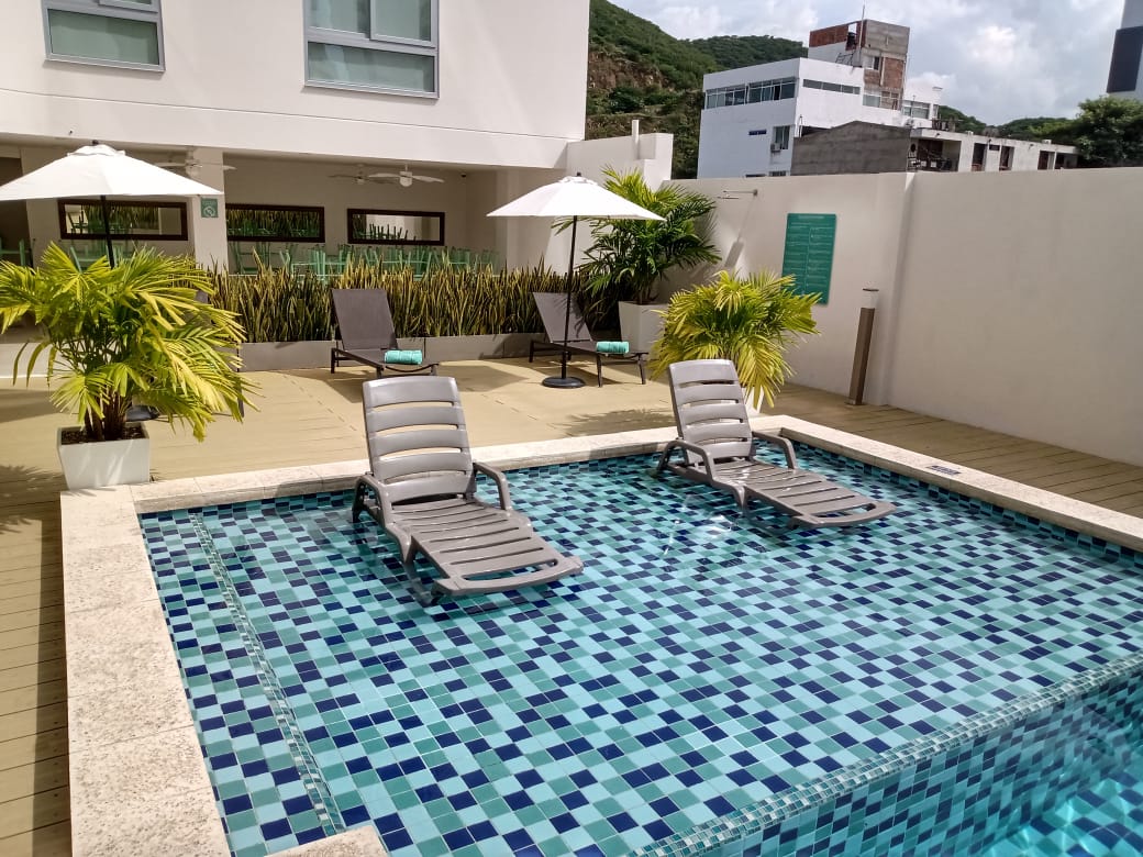Una pequeña piscina con dos tumbonas, con sombrillas blancas y un edificio al fondo.