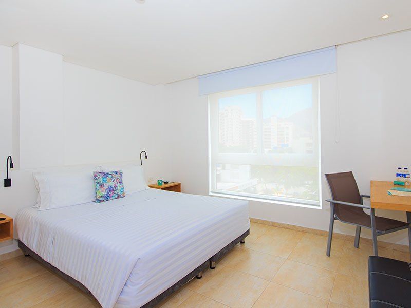 Un dormitorio de hotel blanco brillante con una cama grande, un escritorio y una ventana con vista a la ciudad.