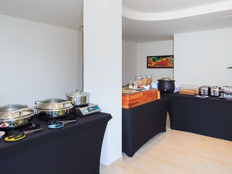 Área de buffet con calentadores de comida y estaciones de servicio sobre manteles negros, bien iluminada con una pared neutra y una pintura de fondo.