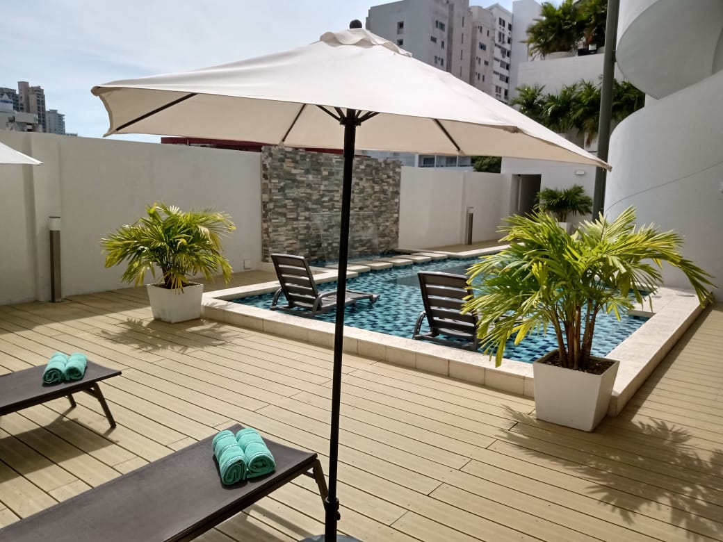 Pequeña área de piscina con sombrilla, tumbonas, plantas en macetas y dos sillas en el área de la piscina, con un edificio de gran altura al fondo.