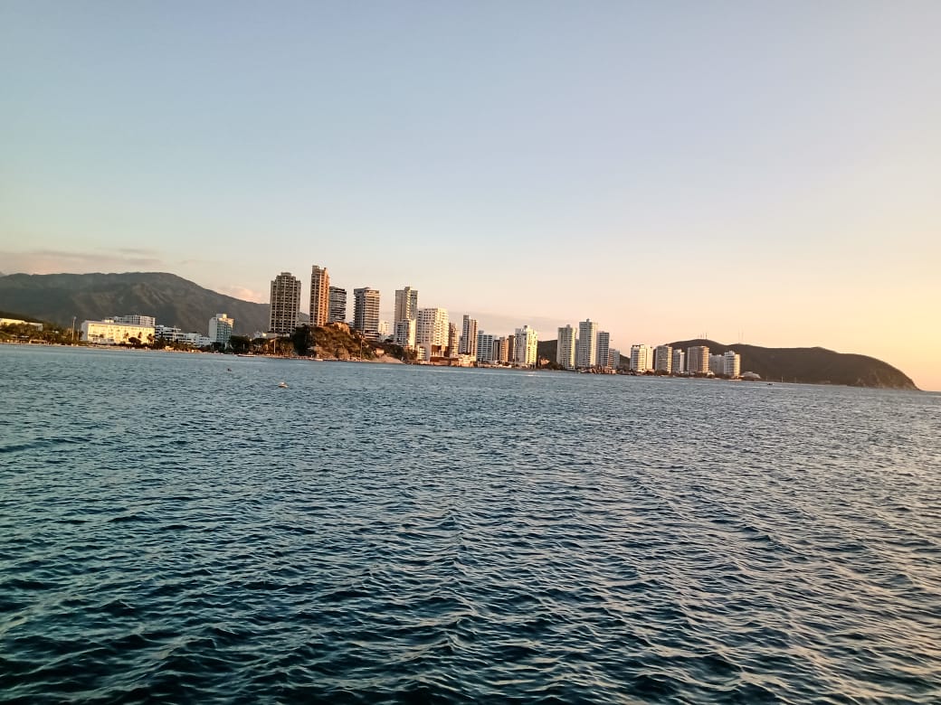 Vista del horizonte de una ciudad a lo largo de una costa al anochecer, con el océano en calma en primer plano y una montaña en el fondo.