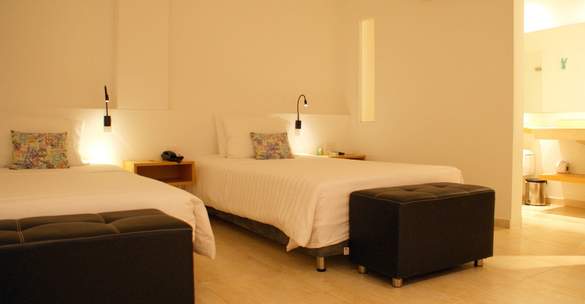 Una habitación de hotel con dos camas individuales, cada una con una pequeña almohada estampada. Cada cama tiene una lámpara de lectura encima. A los pies de cada cama hay una otomana negra.