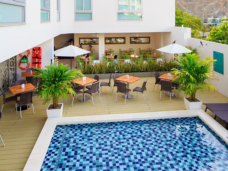 Patio exterior del hotel con mesas, sillas y piscina. Palmeras y sombrillas blancas dan sombra.