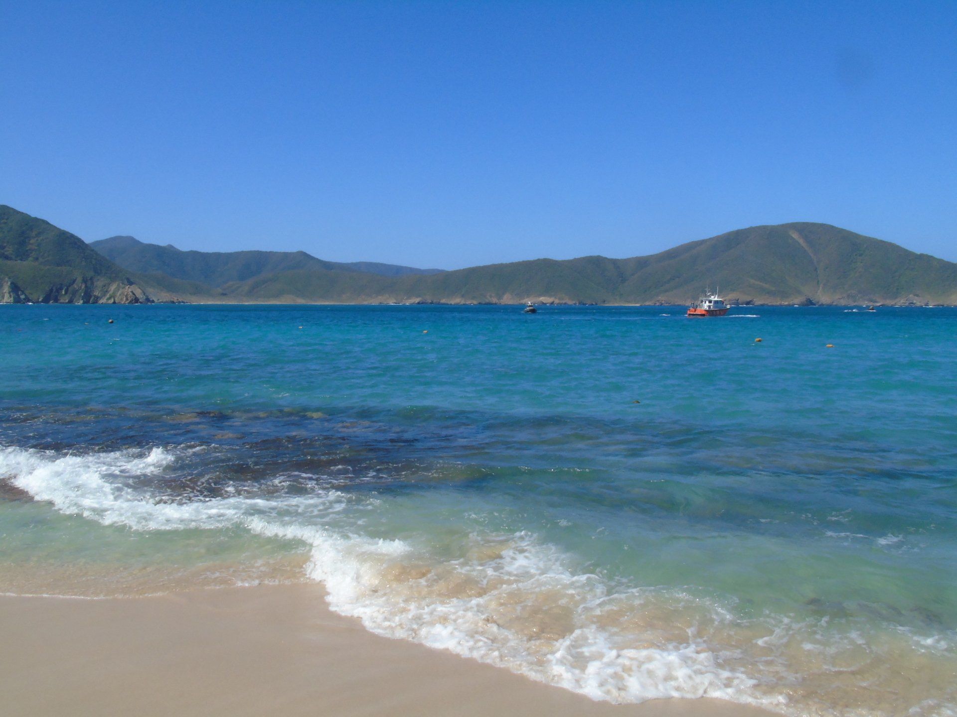 Playa con aguas turquesas, arena blanca y montañas verdes bajo un cielo azul despejado. Se ve un barco a lo lejos.