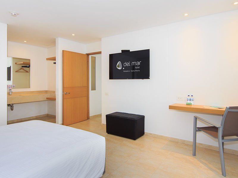 Habitación de hotel con cama, televisor colgado, un pequeño escritorio y baño visible al fondo. La habitación está bien iluminada con tonos neutros.