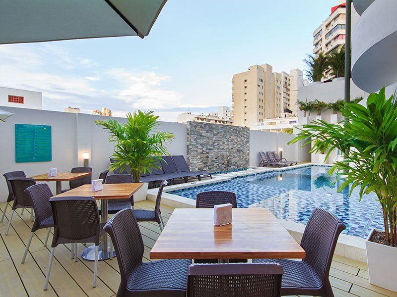 Piscina en la azotea con tumbonas y mesas. Zona de comedor junto a la piscina en un entorno urbano con palmeras.