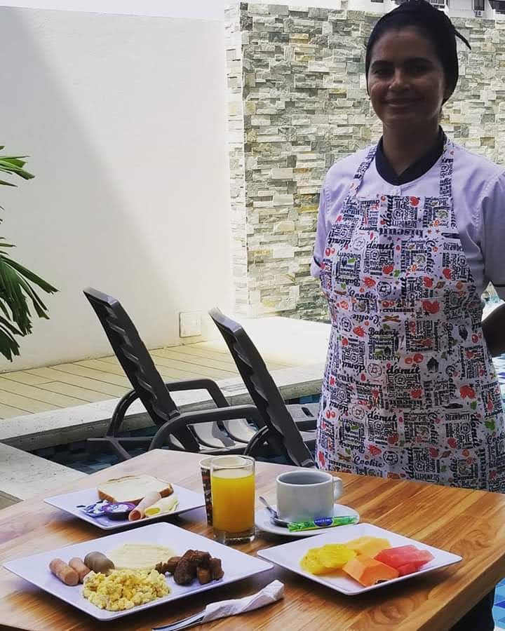 Una mujer con delantal de chef está de pie junto a una mesa preparada para desayunar cerca de una piscina. Los platos incluyen fruta, huevos, salchichas y tostadas.