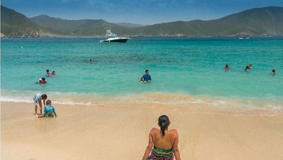 Gente nadando y jugando en aguas turquesas en una playa soleada, con un barco en la distancia y montañas al fondo.