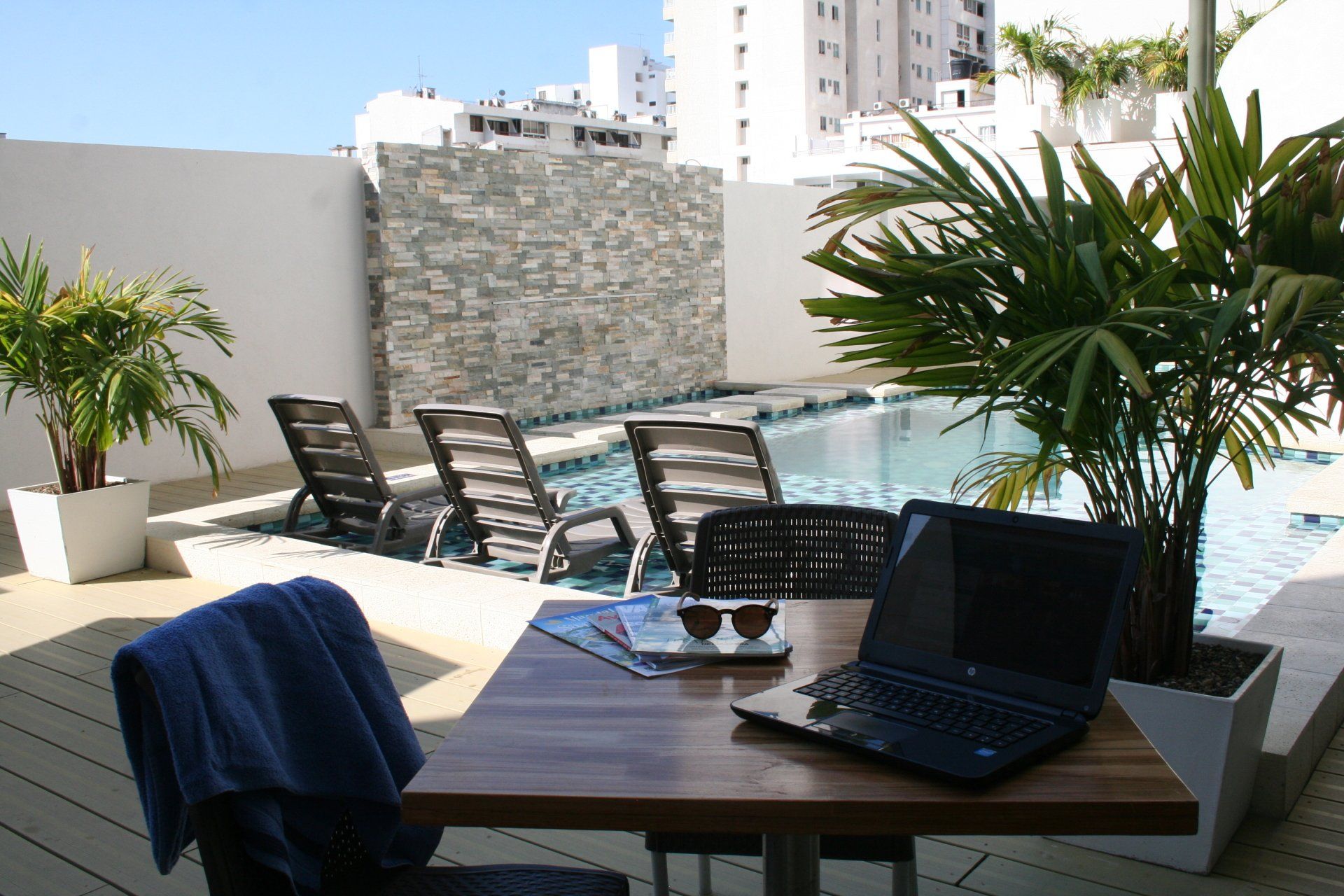 Una computadora portátil y gafas de sol sobre una mesa junto a una piscina con tumbonas, plantas y un muro de piedra; día soleado.