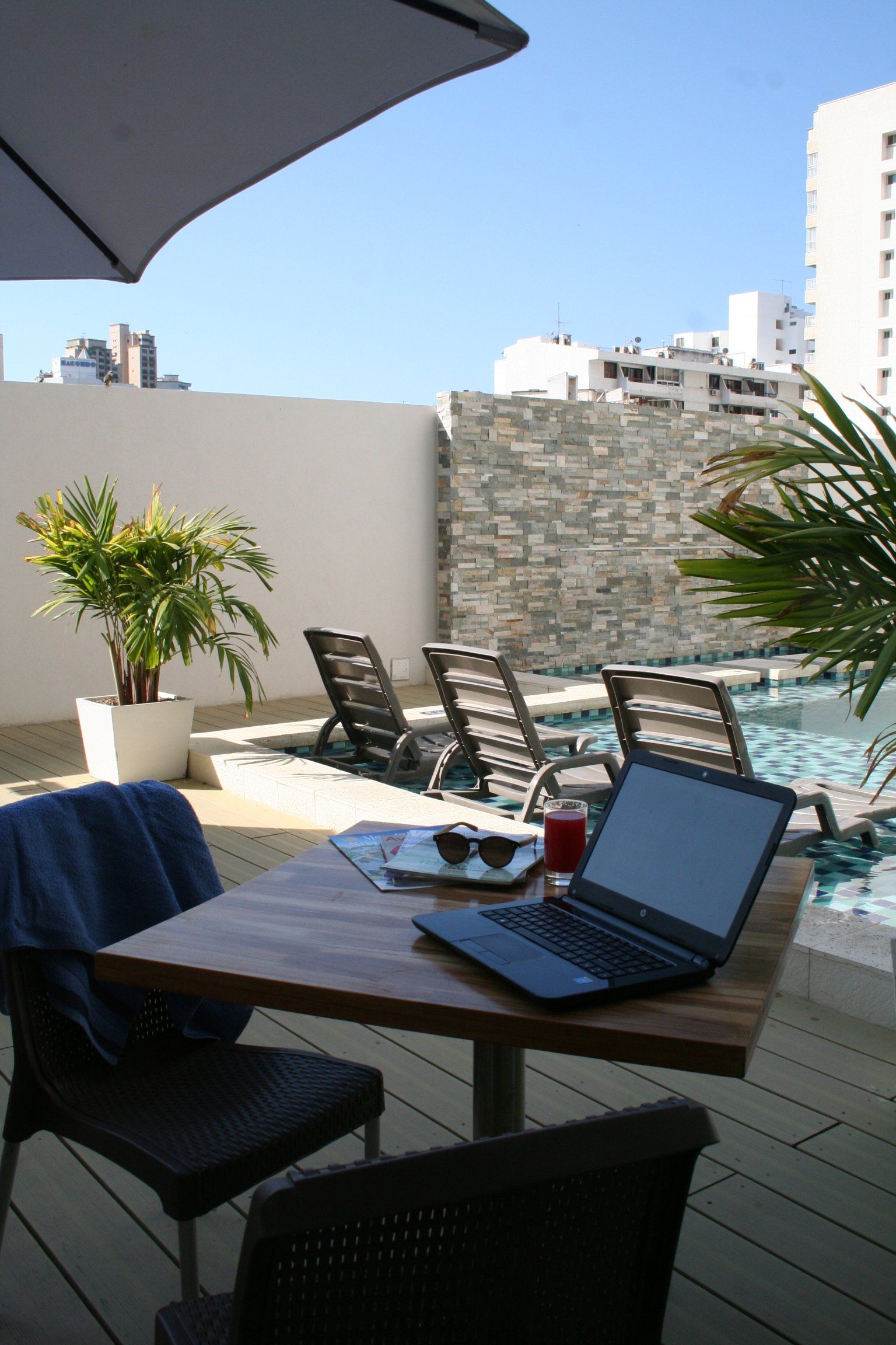 Una computadora portátil y una bebida sobre una mesa junto a una piscina, con tumbonas y edificios de la ciudad de fondo en un día soleado.