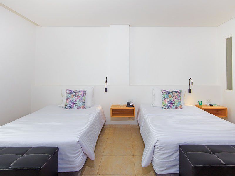 Dos camas individuales con sábanas blancas y cojines florales en una habitación blanca. Cada cama tiene un banco negro a los pies y una mesita de noche de madera.