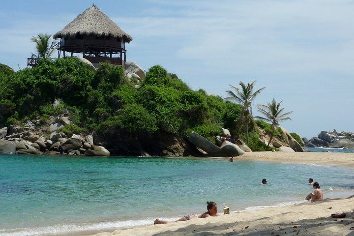 Escena de playa con aguas turquesas y arena blanca. Una estructura con techo de paja se alza sobre un afloramiento rocoso con exuberante vegetación. La gente toma el sol y nada.