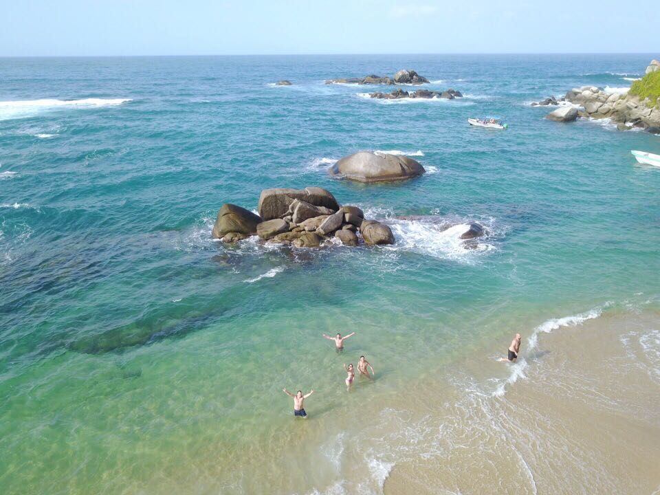 Gente nadando y de pie en una playa de aguas turquesas con rocas en el mar. Nublado, de día.