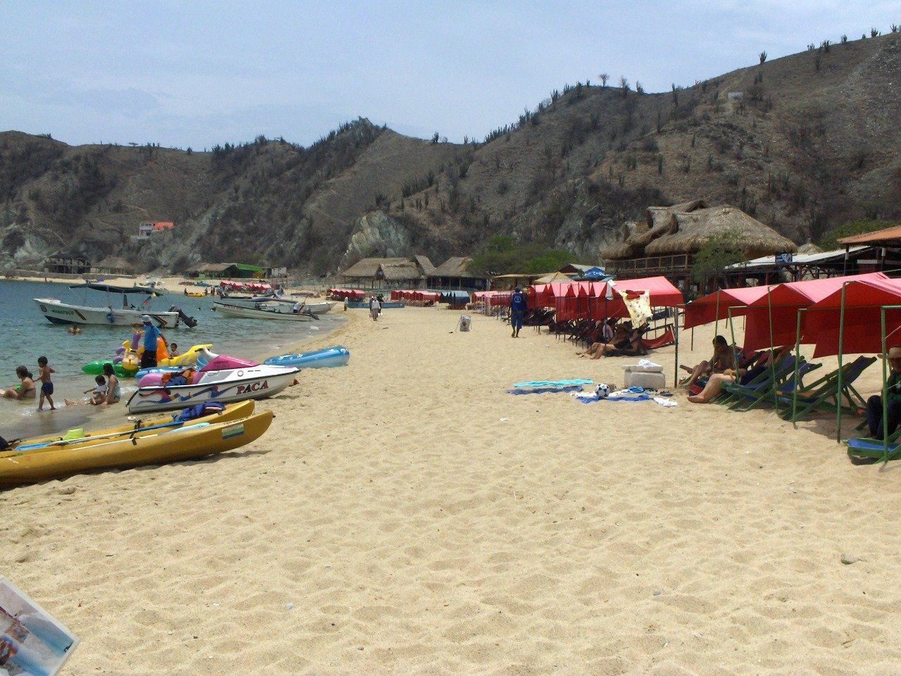 Escena de playa de arena con gente, sombrillas rojas, barcos y colinas al fondo.