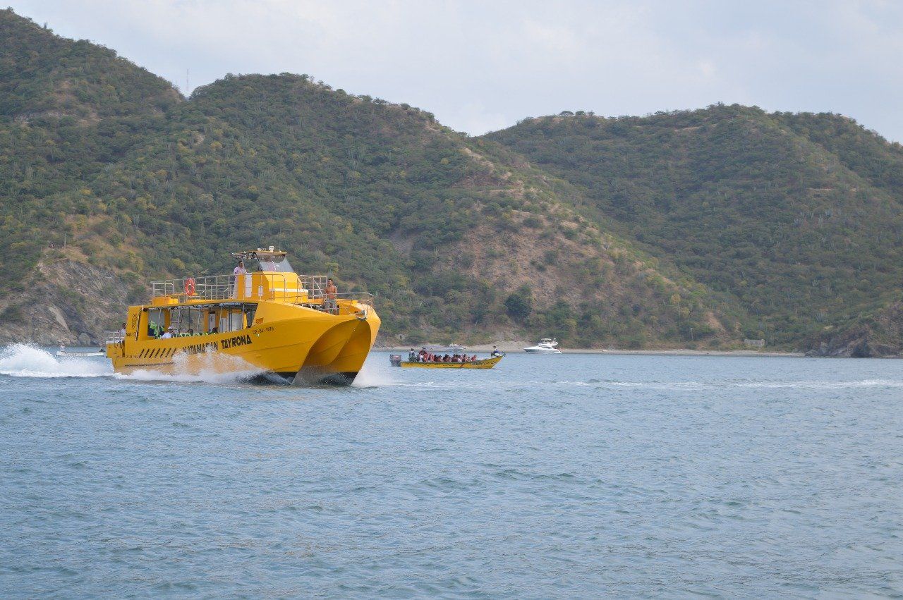 Un catamarán amarillo avanza a toda velocidad por el agua azul, con un pequeño bote al fondo y montañas a lo lejos.