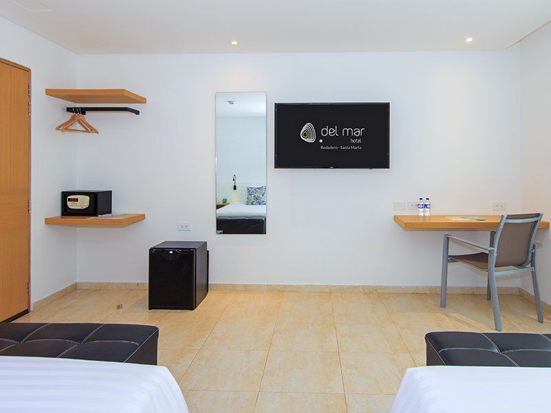 Habitación de hotel con dos camas, televisor, escritorio y una pequeña nevera. Las paredes son blancas y el suelo de baldosas claras.