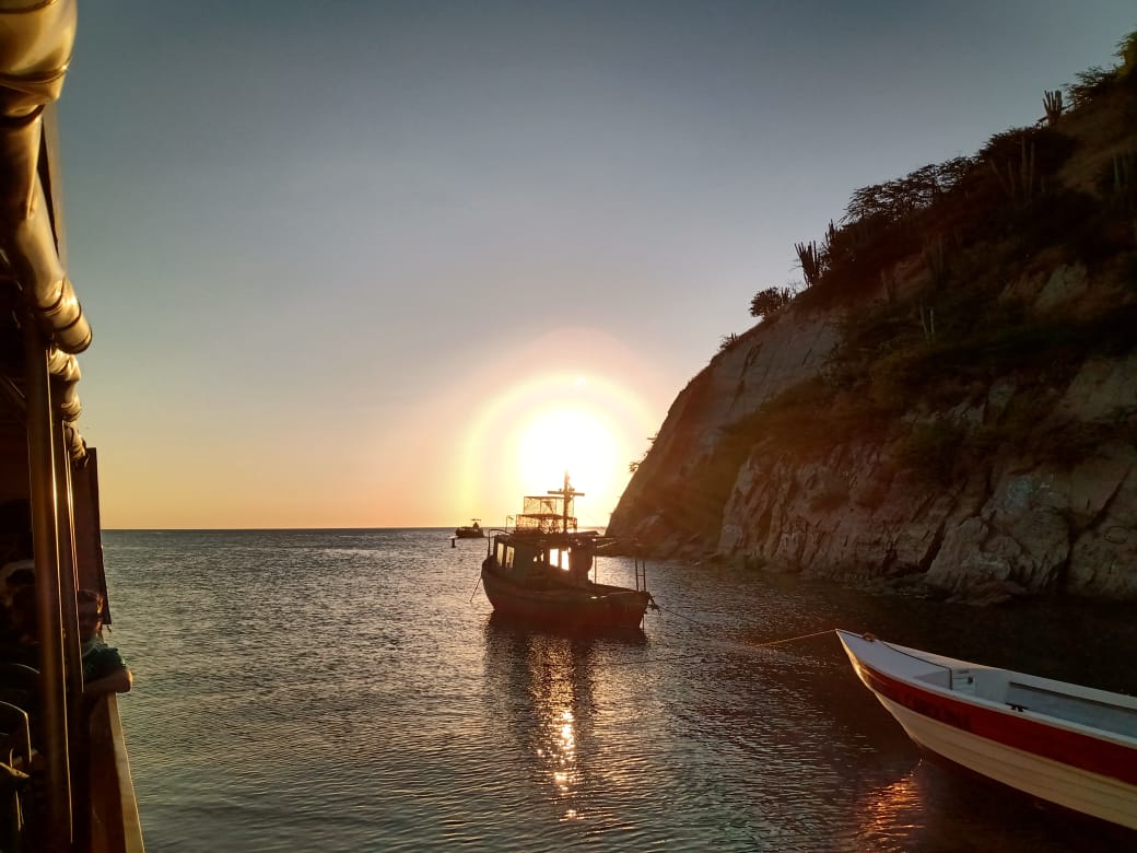 Puesta de sol sobre un océano en calma con dos barcos, uno en la distancia y otro en primer plano, a lo largo de una costa rocosa.