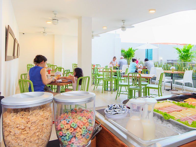Una cafetería luminosa al aire libre con sillas y mesas verdes. Hay gente comiendo; los dispensadores de cereales y la barra de comida están en primer plano.